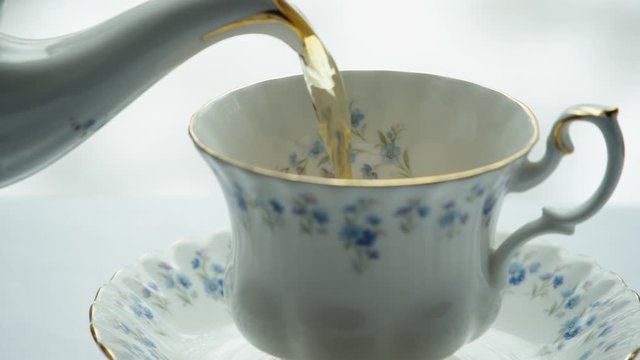 Close-up of tea being poured into a fine china cup on a saucer on a white tablecloth.
