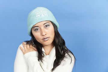 Stunning young Asian woman in winter scene - shot in studio against blue background - wearing thick sweater, cap and with light blue lipstick and eye shadow