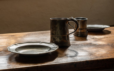 Antique pewter drinking mugs and flat plates with no food on large wooden dining table