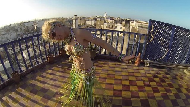Closeup camera circles belly dancer as she spins and does head tosses at sunset on the roof of a building in Morocco.