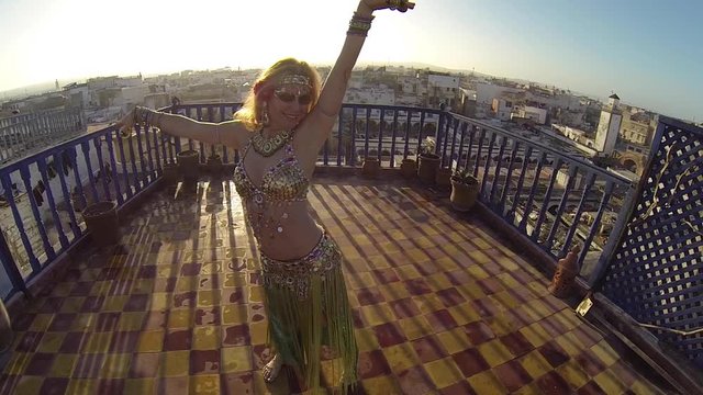 Beautiful blonde belly dancer on the roof of a building at sunset doing snake arms.