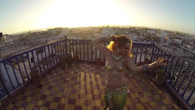 Camera pushes in on beautiful belly dancer as she does hip drops on the roof of a building in Morocco at sunset.