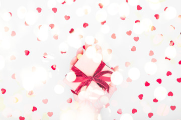 Craft box with red ribbon bow in female hand. Valentine day and eco-friendly wrapping concept. Trendy minimalistic flat lay design background. Horizontal with festive bokeh lights