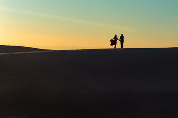 Fototapeta na wymiar A couple on a desert dune in silhouette at sunset with the woman holding her dress.