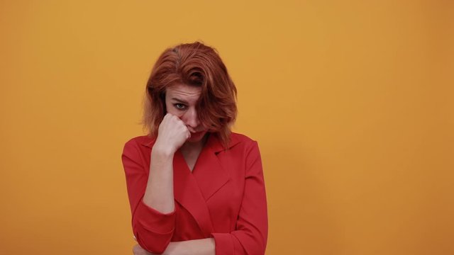 Boring caucasian woman looking at camera, pointing fist aon cheek, keeping hand on stomach wearing fashion red jacket over isolated orange background. People lifestyle concept.