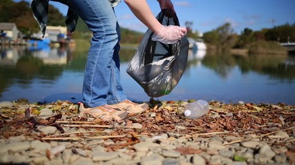Concept of cleaning the environment. A woman walks along the river Bank and collects garbage and bottles in a bag. River on the background