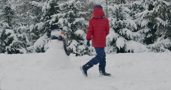 Caucasian boy destroys funny snowman dressed in stylish hat and black scarf in winter