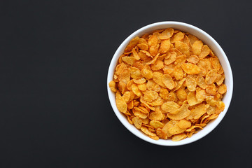 Dry corn flakes in a white bowl on a black metallic surface. Copy space.