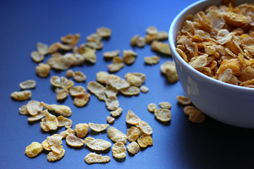 Corn flakes, dry, in a white bowl on a bluish black metallic surface.