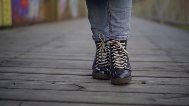 Close-up of women's legs in jeans and black shoes with lace-up heels, they are walking on a wooden bridge, in the background a wall with graffiti.