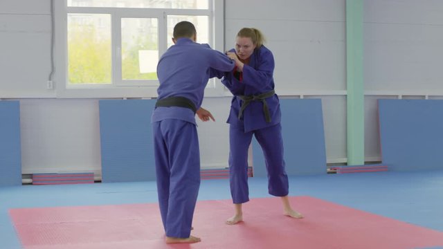 Arc shot of young barefoot woman in jujitsu gi practicing fighting technique with male partner in gym