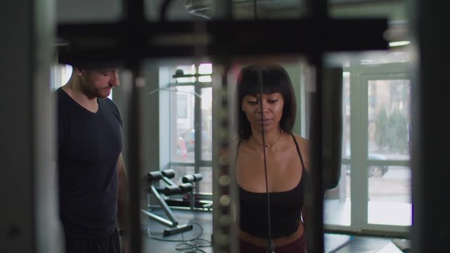Determined african american fit woman exercising cable rope triceps extension during upper-body workout routine in fitness club with fitness instructor. Female doing rope pushdowns for triceps at gym.