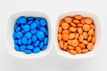 Two squared small bowls with small orange and blue sugar coated chocolate candies isolated on white background, top view or flat lay photograph of unhealthy sweets