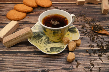 A Cup of black tea, tea leaves, pieces of brown sugar, oatmeal cookies, waffles on a wooden background. Close up.