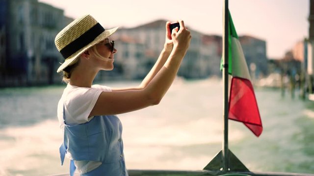 Positive hipster girl in stylish hat enjoying time for recreating during italian weekend taking video of architecture using camera on modern mobile phone while floating on touristic boat with flag