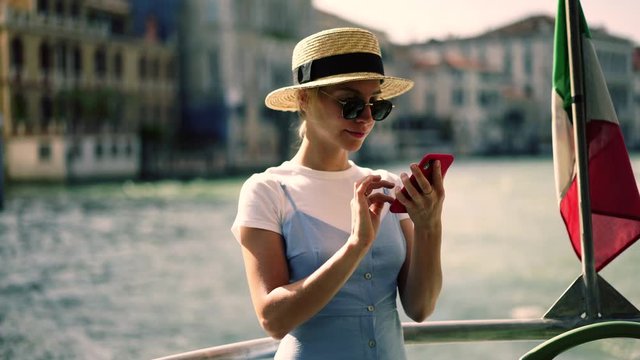 Cheerful female tourist in stylish dress making cellphone images of architecture buildings during boat excursion around Venice city, positive smiling hipster girl enjoying summer vacations in Italy