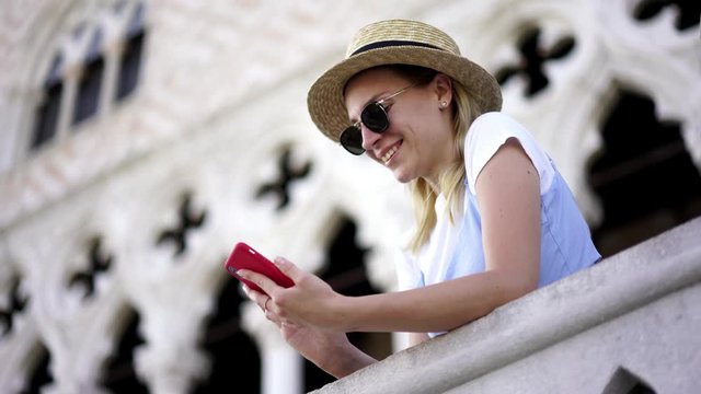 Happy woman in hat and glasses searching information about sightseeing in Venice while messaging with followers in social network. Smiling hipster girl connected to 4g roaming internet on mobile phone