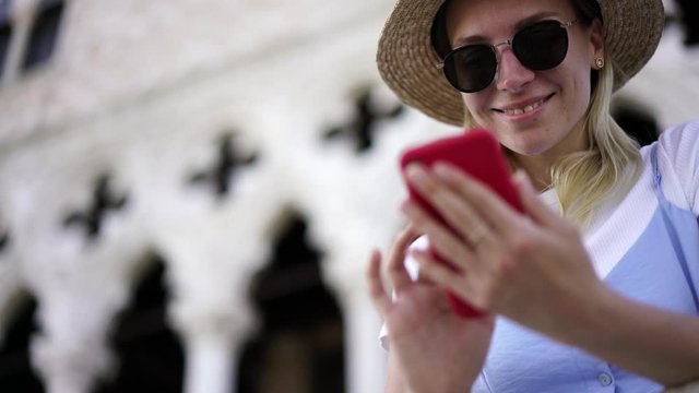 Happy cheerful young woman in hat and sunglasses messaging with followers in social network connected to 4g roaming internet on mobile phone, positive hipster girl enjoying vacation holidays in Italy