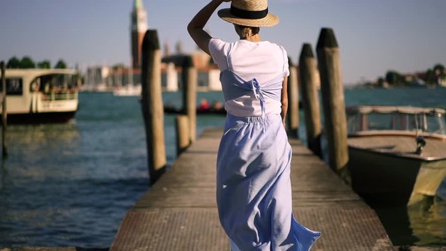 Back view of millennial female tourist running on Venice pier while summer wind blowing stylish blue sundress, woman traveller making photos for website via modern cellular phone during trip to Italy