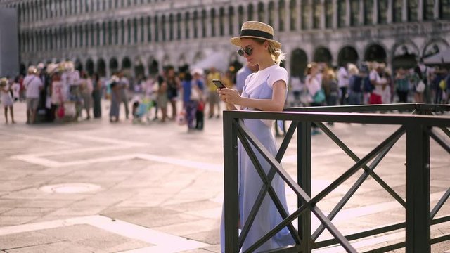 Positive cheerful female tourist using mobile phone application for communicate with people on travel messaging about sightseeing in Venice while resting and smiling on St Mark's Square. Using tech