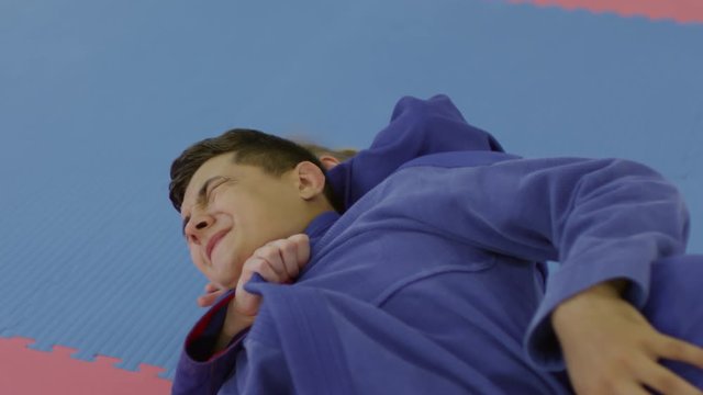 Slow motion shot of young male jujutsu athlete wincing in pain while being choked by female sparring partner during training fight on mat in martial arts gym