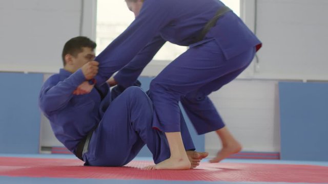 Sequence of shots of male martial arts athlete in blue uniform sitting on floor, pulling and throwing female partner down on mat and doing chokehold during jujutsu practice