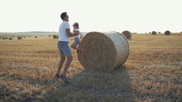 Little daughter jumping on father's arms from a big haycock in field