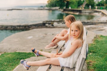 Little girls playing by the lake in summertime, children having fun outside, resting on the bench