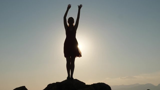 Young positive woman dancing silhouette, sun light flash from behind when she wriggle body. Girl stand on stone at sea beach, raise hands and wave in air, bend and move body in rhythm