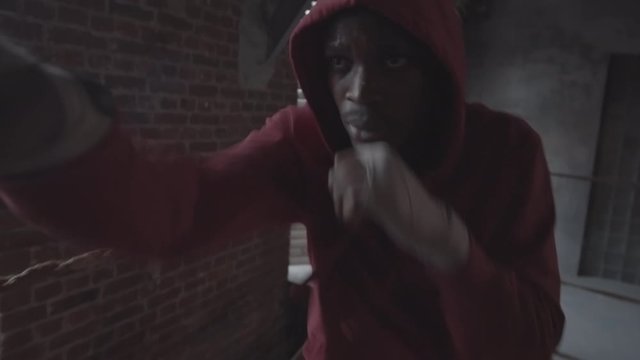Waist-up shot of young Afro-American man in hooded sweatshirt shadow boxing in ring at fight club in dingy industrial warehouse, delivering vigorous punches, weaving and ducking