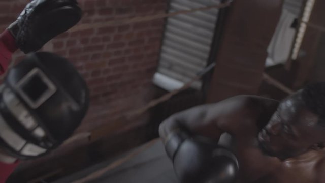 Medium high-angle shot of focused Afro-American boxer with bare chest and gloves practicing jabs on punch mitts while training with coach in dimly lit boxing gym
