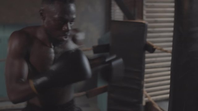 Waist-up handheld shot of young African American boxer in shorts, with bare torso and gloves furiously hitting heavy punch bag with forceful blows in rundown underground fight club