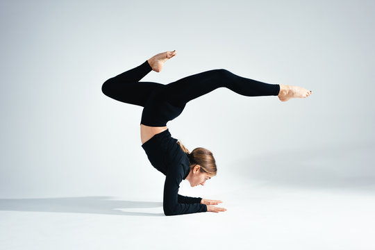 Young Handsome Fit Woman Wearing Black Sportswear Doing Stretching Body Against White Background. Studio Shot. Flexible Girl Doing Yoga Poses In White Room