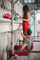 Two workers stands high on the scaffolding, plastering and renovating the wall on the old utility building	