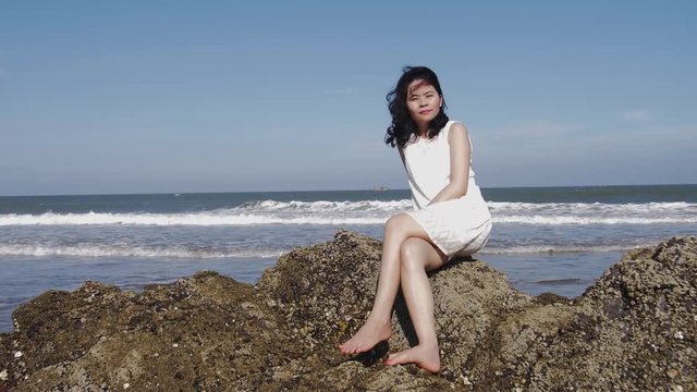 Dark-haired woman looks toward camera while sitting on rock by ocean in Vietnam, slomo