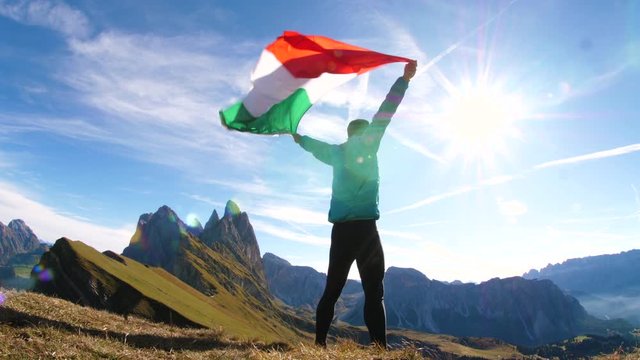 Young man in running clothes standing on mountain peak raising italian flag to the sky in windy sunny day. Seceda, Puez Odle, Dolomites, Italy.