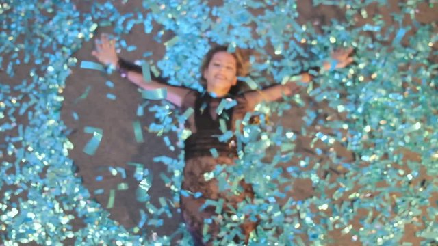 Crazy New Year's Eve. A beautiful lady lies on the floor and creates a snow angel out of blue confetti in slowmo
