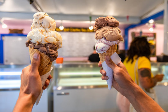 A close up and first person perspective of a man holding two waffles cone with scoops of ice cream at a shop, with blurry menu in the background