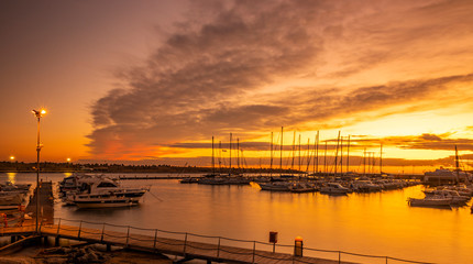 Obraz premium A view of the marina of Santa Maria di Leuca at red and orange sunset. Sailboats and yachts moored on the docks of the port. Wooden walkways lead to harbor quay. Puglia, Italy, Lecce, Salento.
