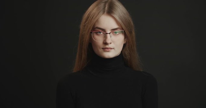 Attractive young woman in eyewear and black turtleneck sweater looking to camera isolated on dark background