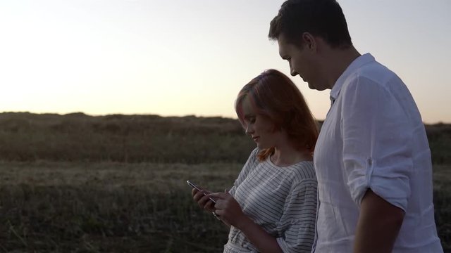 Closeup of romantic couple in love are standing and watching in smartphone. Man and woman are at the green field. They are stanging close to each other and the sky is bright.