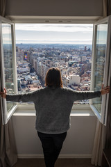 Obraz premium rear view of young woman opening a window from her room and looking outside on a sunny day with Madrid skyline views