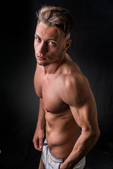 Fototapeta premium Handsome shirtless athletic young man wearing only pants, with blue eyes, looking at camera in studio shot, on dark background