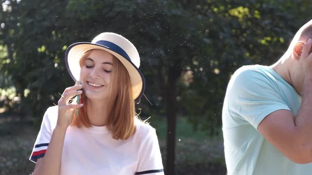 Portrait of pretty positive teenage girl with red hair wearing straw hat talking happily on mobile phone while her boyfrient is waiting bored beside.