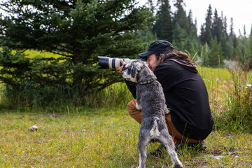 Young male photographer kneeling down to take a picture with a zoom lens, and small grey dog...