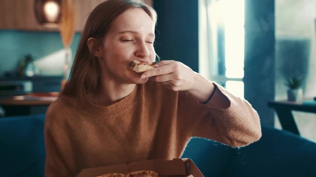 Cheereful european young woman in brown sweater sitting on sofa at modern living room and greedily eating tasty pizza, enjoying and smiling. Fast food addiction. Slow motion.