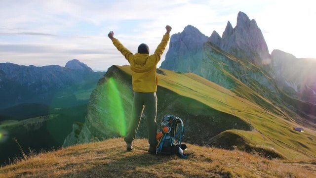 Young man hiking at Seceda mountain raising arms to the sky on the peak at sunrise. Backpack, yellow jacket, boots, beanie. Traveling to puez Odle, Dolomites, Trentino, Italy.