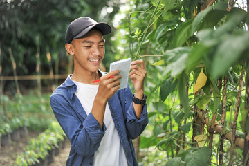 Fototapeta premium Portrait of smiling young Asian farmer man checking quality with small notebook. Happy young Asian farmer at the garden