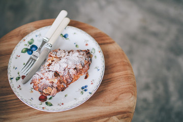 Almond croissants topped with icing sugar on white plate with fork and knife wooden table with female hands holding fork and knife woman holding and eating a piece of croissant