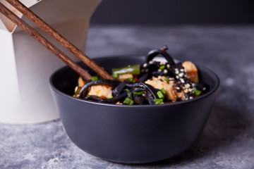 Black squid ink udon noodles with chicken meat on the black bowl with chopsticks on dark stone background and paper box nearby. Take out food concept.